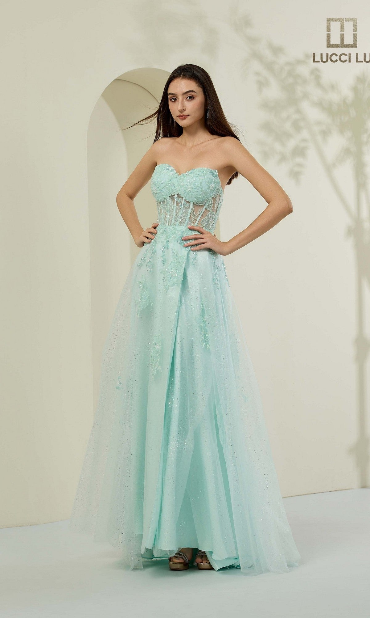 A woman poses in the Lucci Lu 1476 long prom dress in mint, featuring floral appliqués and a sheer strapless corset bodice, against a neutral background.